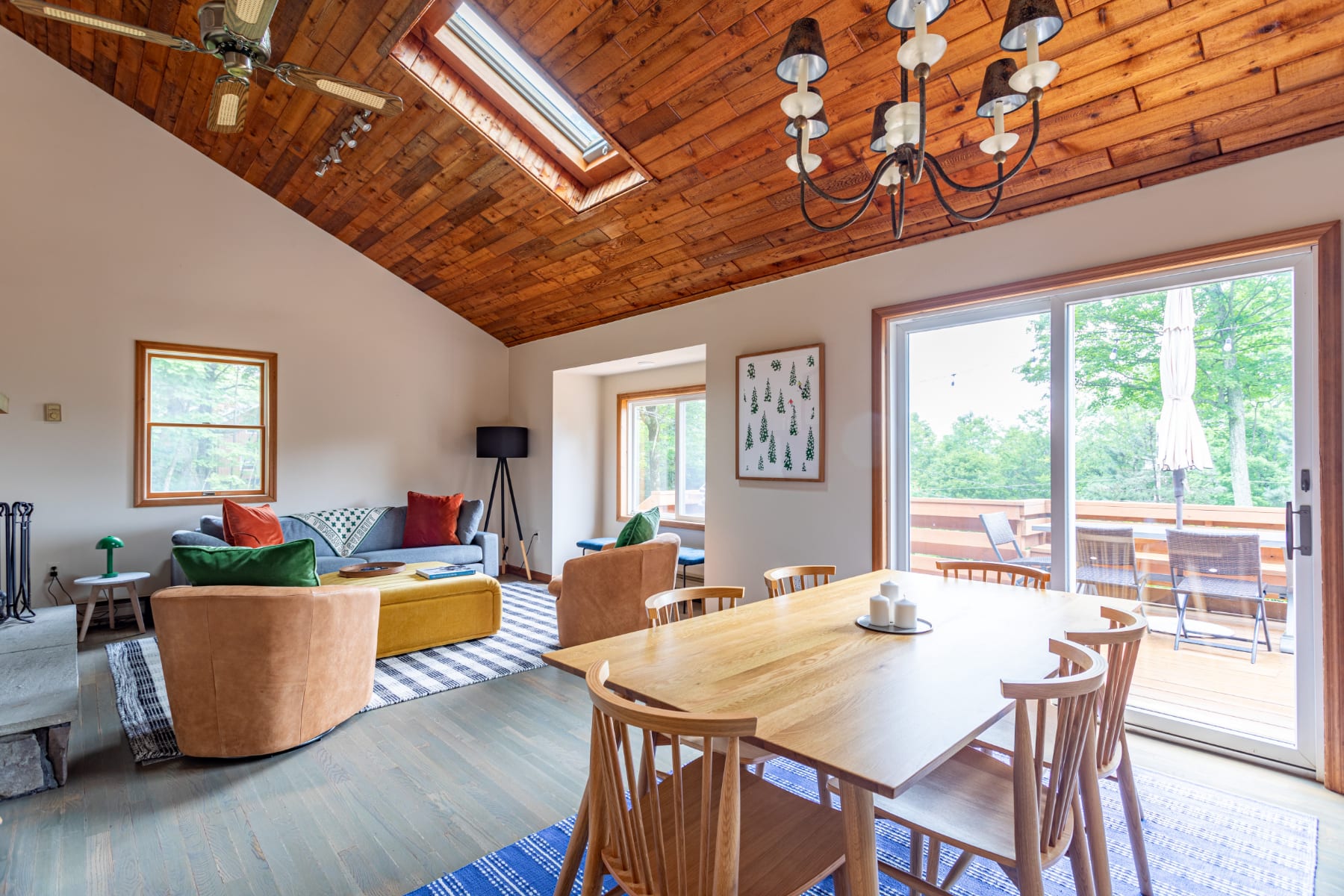 Spacious dining area with a light wood table and chairs, adjacent to a living room with a gray sofa and accent chairs, all beneath a vaulted wood-paneled ceiling with a skylight.
