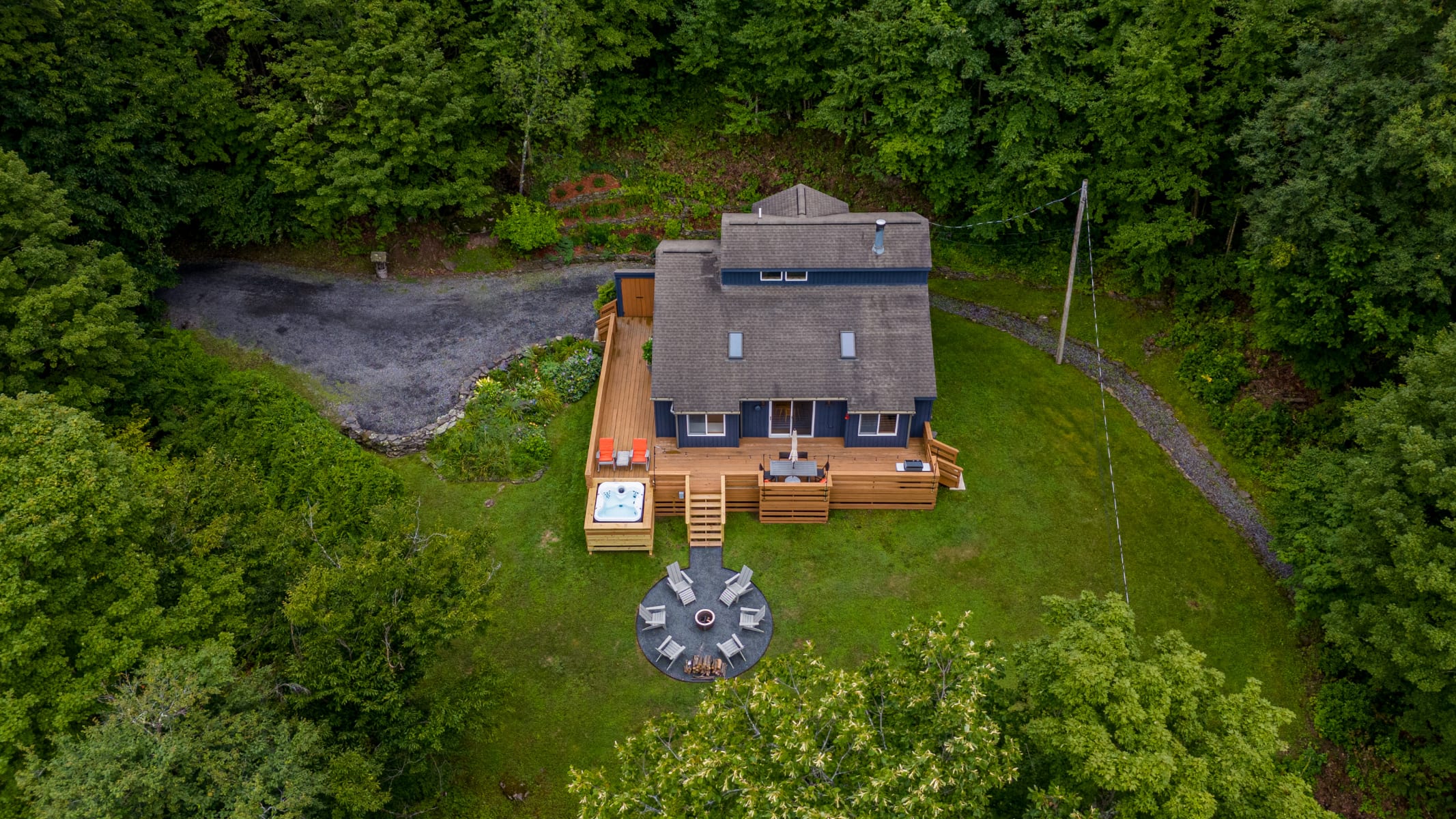 A high aerial view of the property, showcasing the dark-sided house with its extensive wooden deck, hot tub, and circular fire pit area. The house is nestled amidst dense green trees, with a driveway and surrounding natural landscape visible.