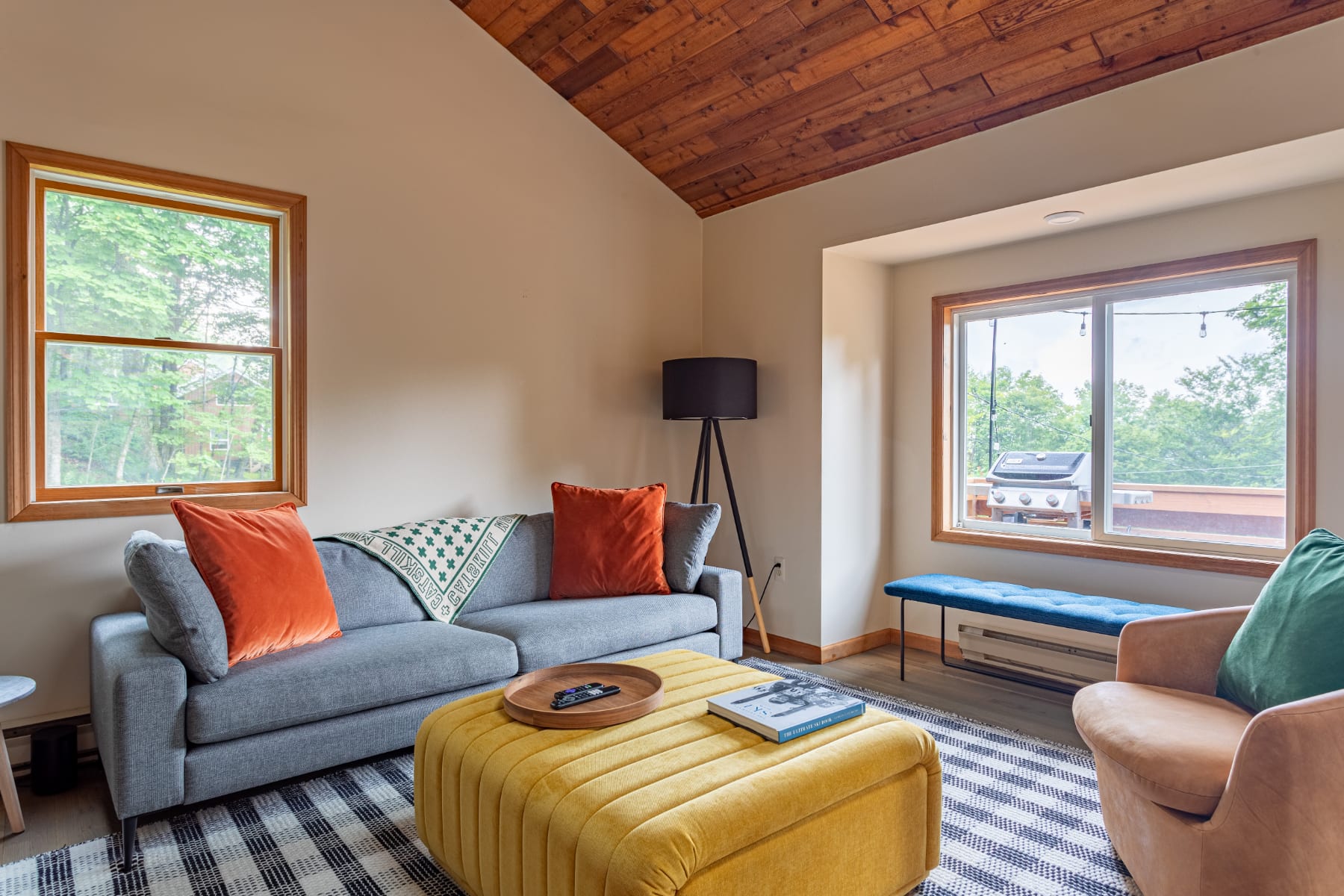 Cozy living room with a gray sofa adorned with orange pillows and a green throw, a large yellow tufted ottoman, and a black and white striped rug, under a vaulted wood-paneled ceiling.