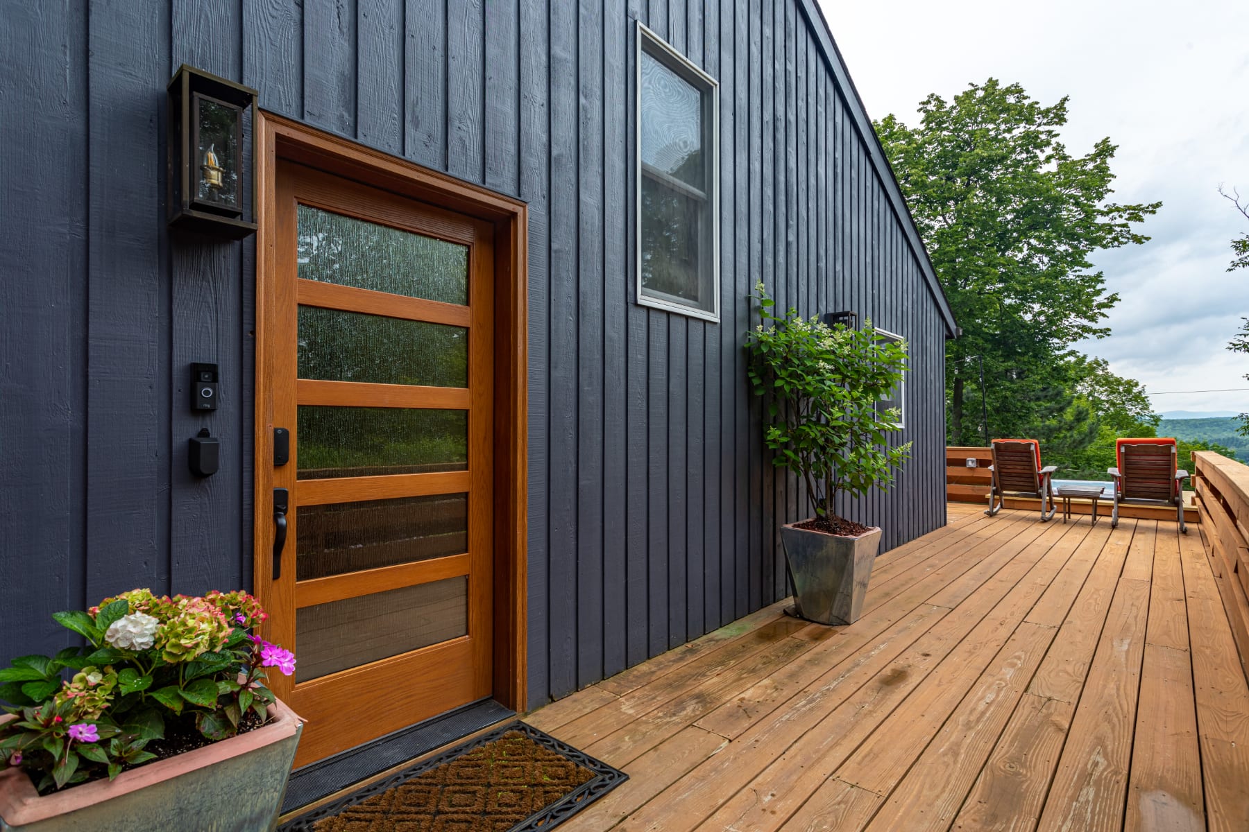 A close-up of the modern dark-sided house entrance, featuring a warm wooden door with glass panels, a contemporary outdoor light, and potted plants. A wide wooden deck extends from the entrance, with distant views of the surrounding landscape.