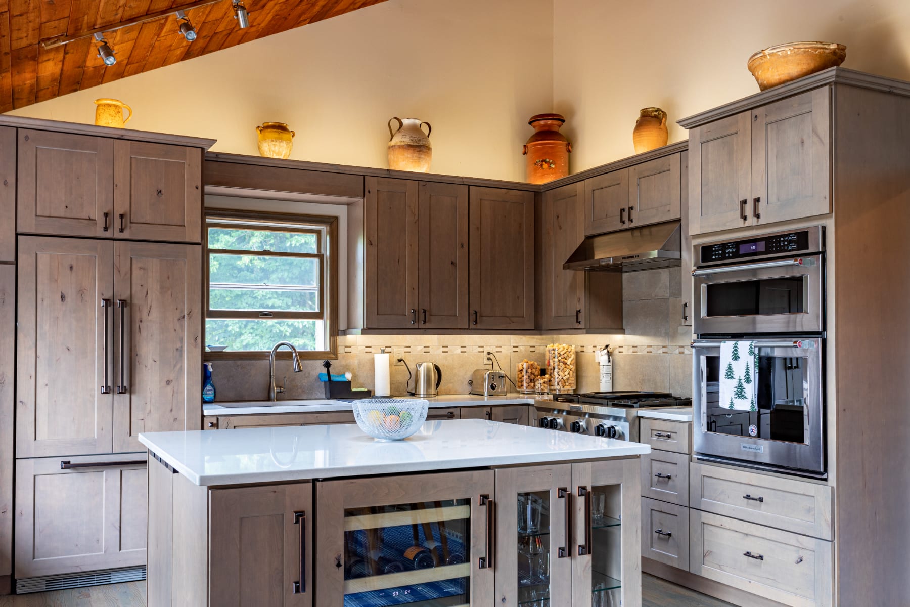 Well-equipped kitchen featuring light wood cabinets, stainless steel appliances including a double oven and range hood, white countertops, and a central island with a sink and wine fridge.