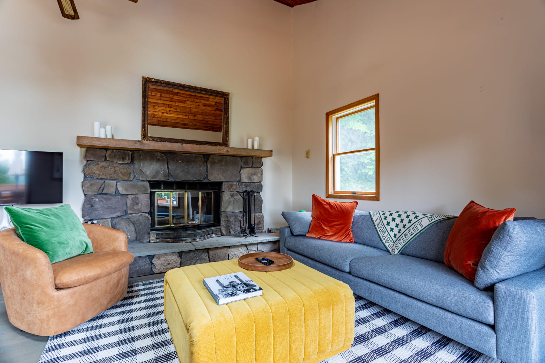 Inviting living room centered around a large stone fireplace with a wooden mantel, flanked by a gray sofa and a tan armchair, with a yellow ottoman on a striped rug.