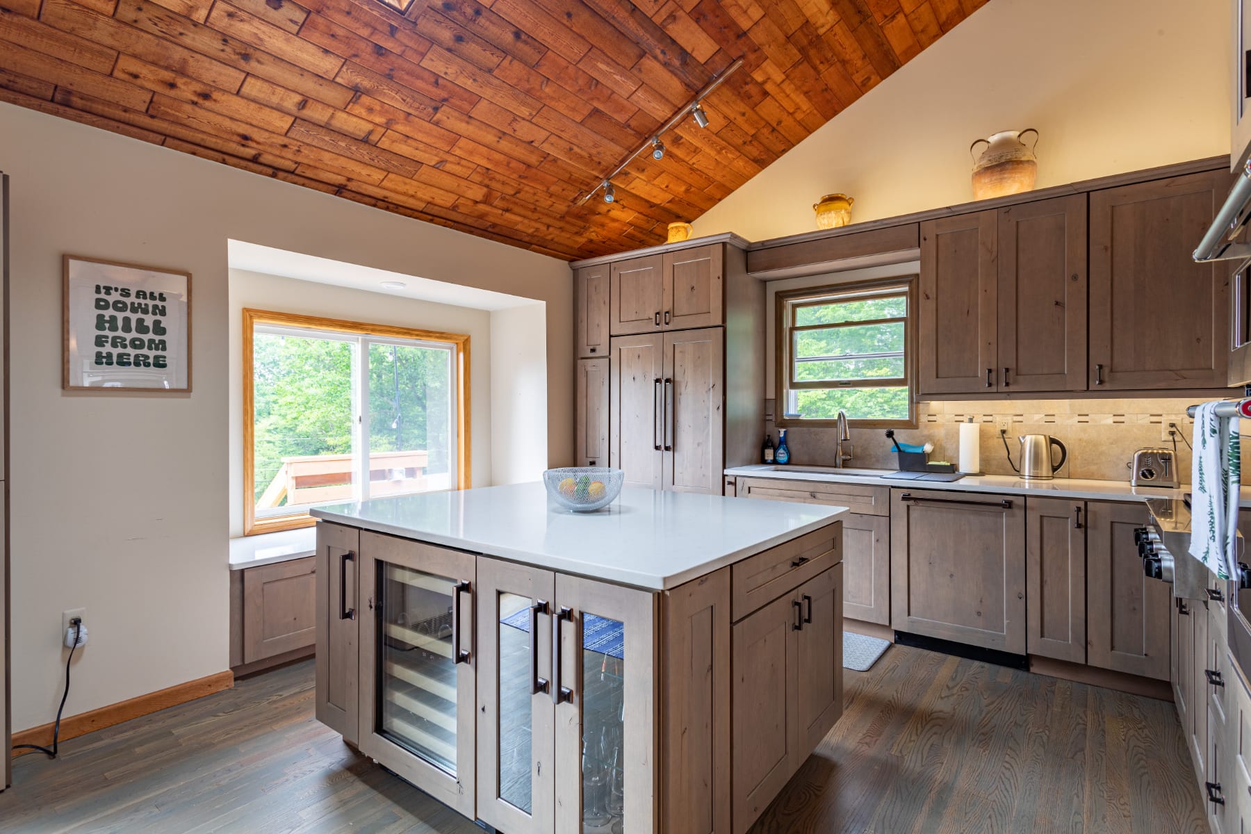 Modern kitchen with light wood cabinetry, white countertops, a large island with a wine fridge and seating, and a vaulted wood-paneled ceiling, illuminated by track lighting.