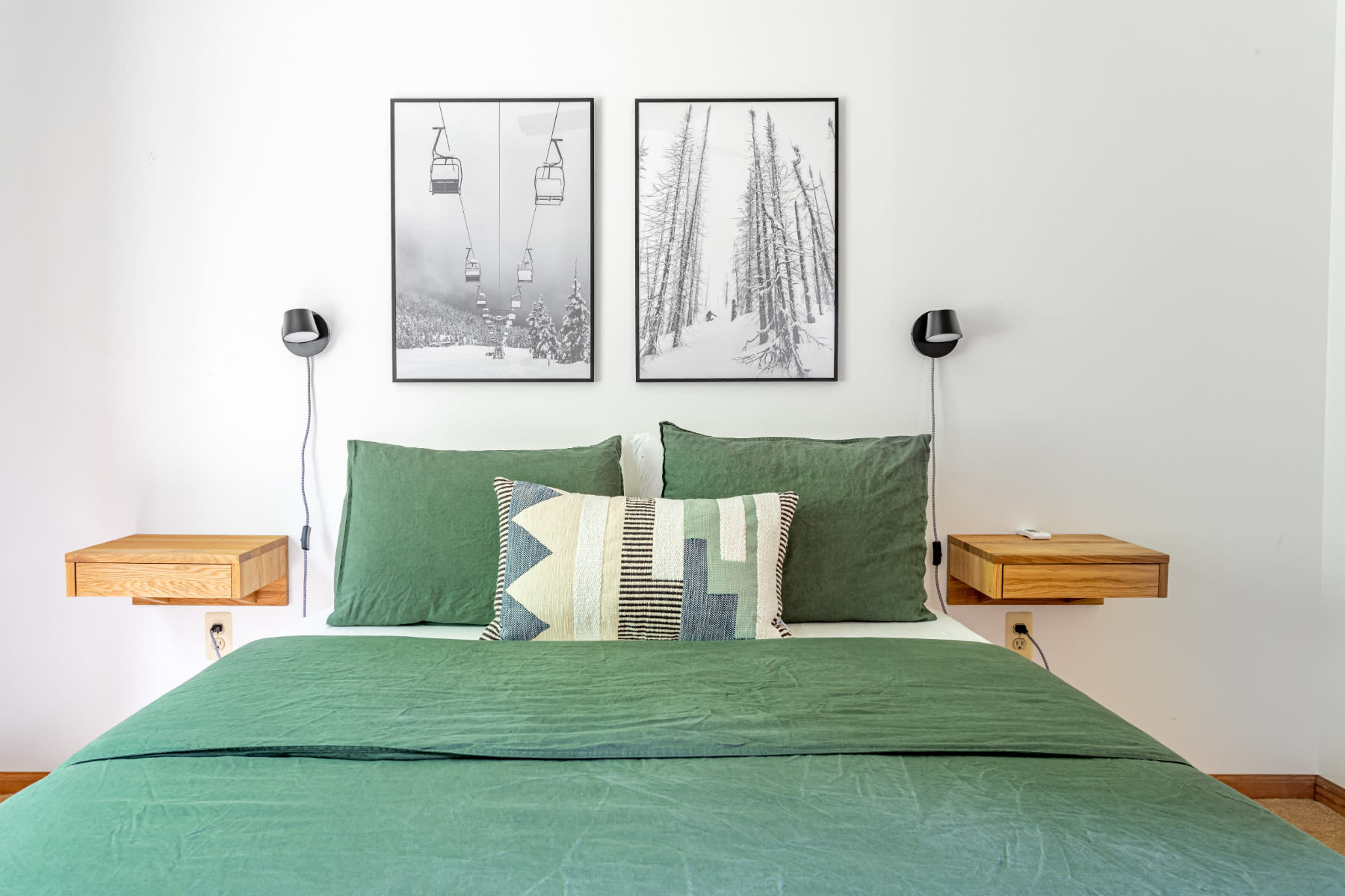 A cozy bedroom featuring a large bed with green linen bedding, flanked by floating wooden nightstands and black wall-mounted lamps. Two framed black and white ski-themed prints adorn the white wall above the bed.