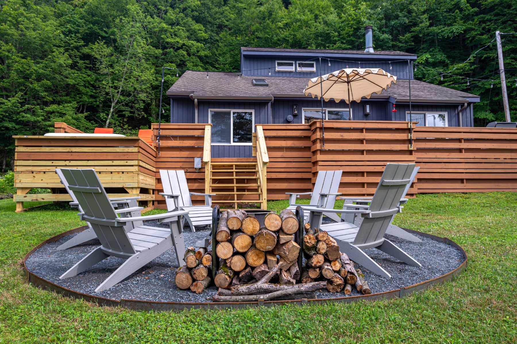 An aerial view of the outdoor fire pit area, showing the circular gravel base with Adirondack chairs and a large stack of firewood. The dark-sided house with its wooden deck and surrounding trees is visible in the background.