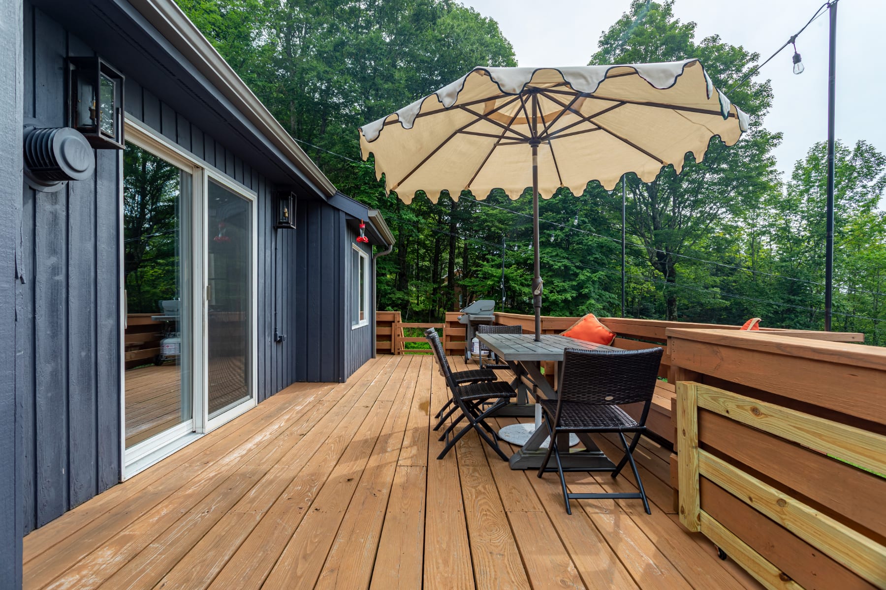 A wide shot of the wooden deck, showcasing the dark-sided house with large sliding glass doors leading to the interior. An outdoor dining set with an umbrella is visible, surrounded by a wooden railing and lush trees.