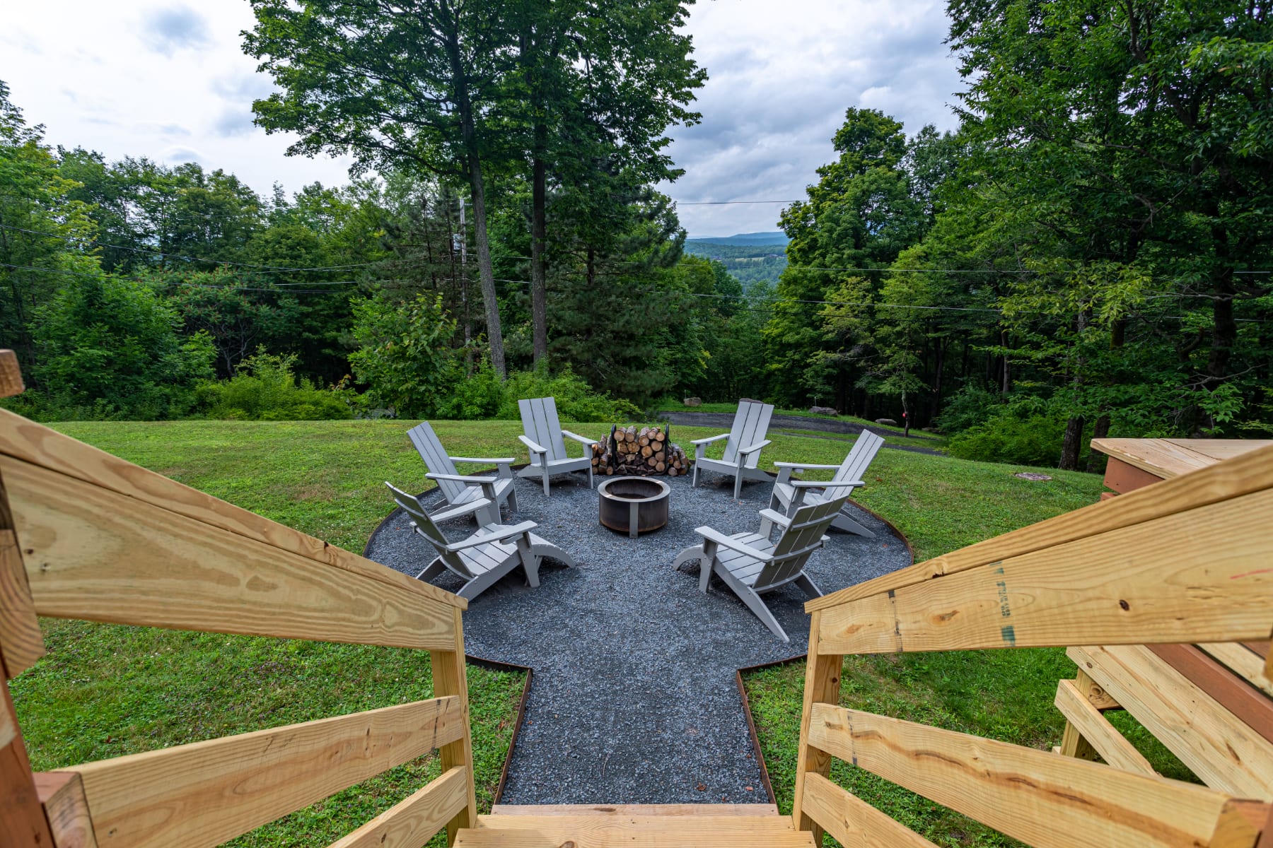 An inviting outdoor fire pit area with a circular gravel base, surrounded by several light gray Adirondack chairs. A stack of firewood is ready for use, set against a backdrop of green grass and dense trees.
