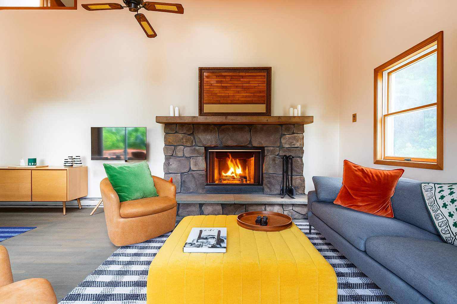 Cozy living room with a large stone fireplace, a gray sofa with orange pillows, an orange armchair with a green pillow, and a yellow ottoman coffee table on a striped rug. A TV is mounted on the wall to the left.