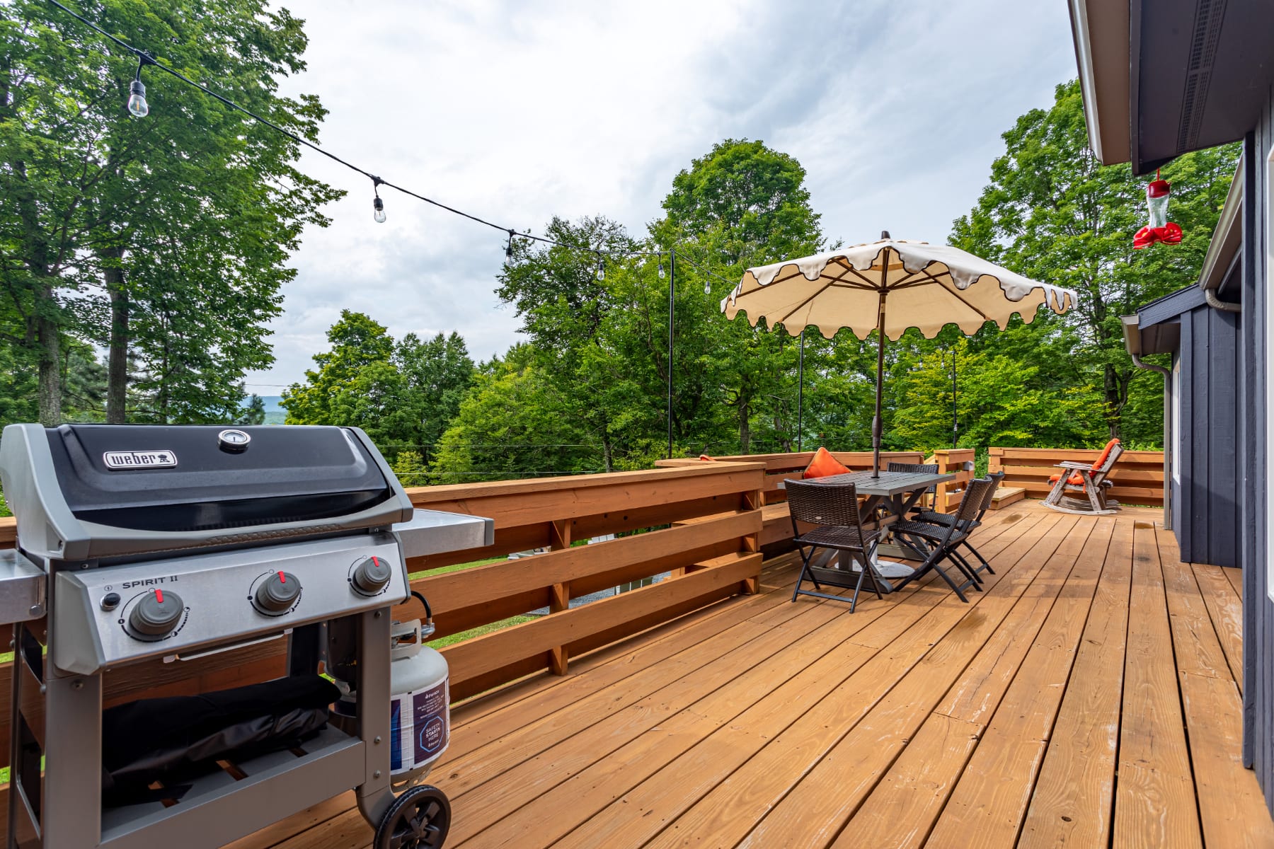 A spacious wooden deck featuring a Weber gas grill and an outdoor dining set with an umbrella, surrounded by lush green trees. String lights are strung overhead, creating an inviting outdoor entertaining space.