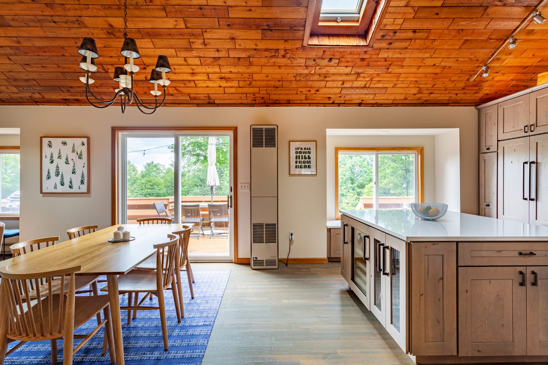 Bright dining area with a wooden table and chairs on a blue rug, leading into a modern kitchen. The space features a vaulted wooden plank ceiling with a skylight and a sliding glass door opening to an outdoor deck.