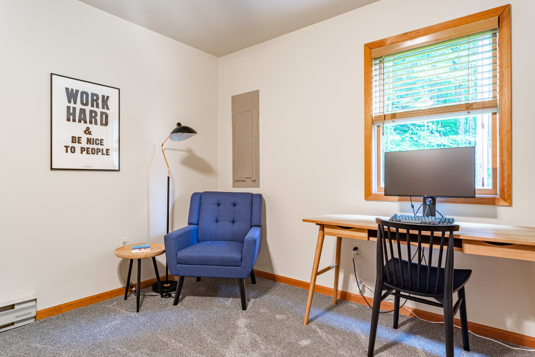 Dedicated office space with a wooden desk and black chair, a blue armchair, a small round side table, and a "Work Hard & Be Nice To People" poster on the wall.