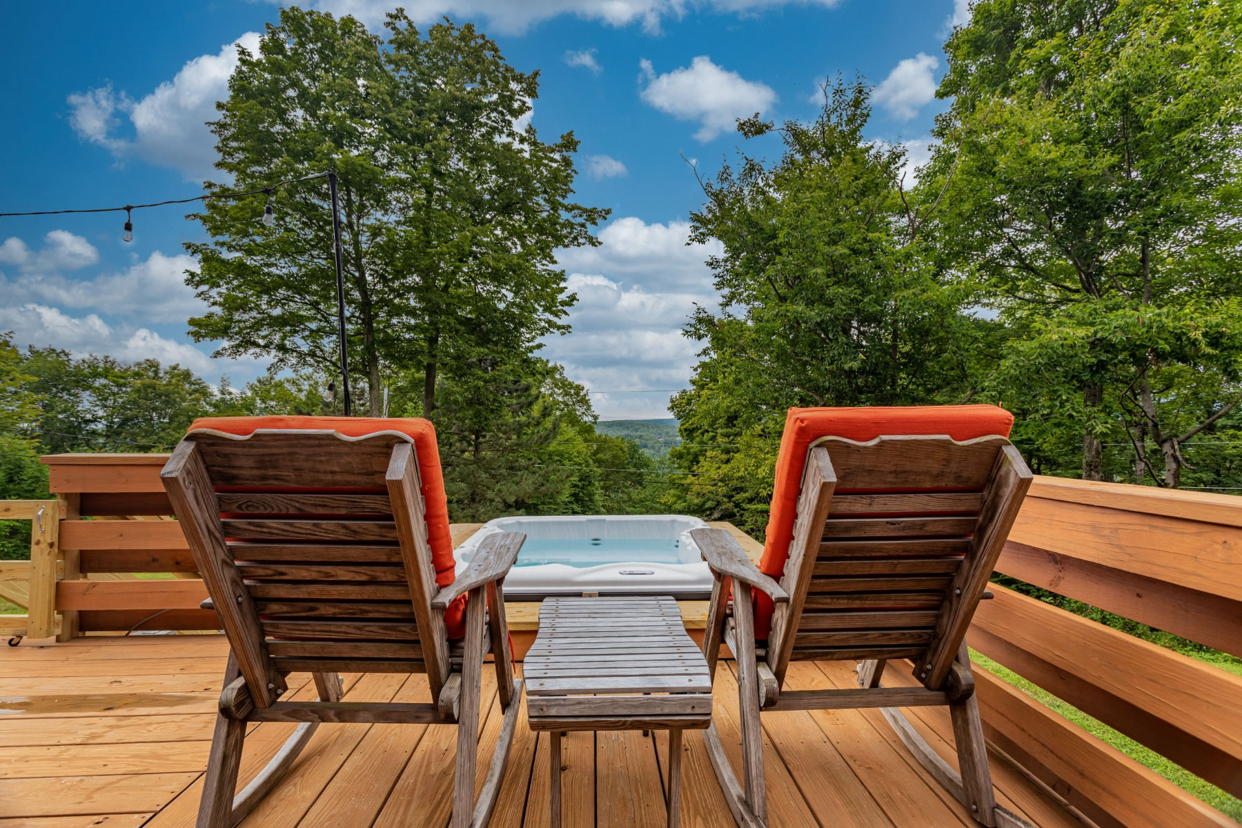 A relaxing hot tub area on a wooden deck, offering scenic views of distant mountains and lush greenery. Two wooden Adirondack-style chairs with orange cushions face the view, with a small table between them.