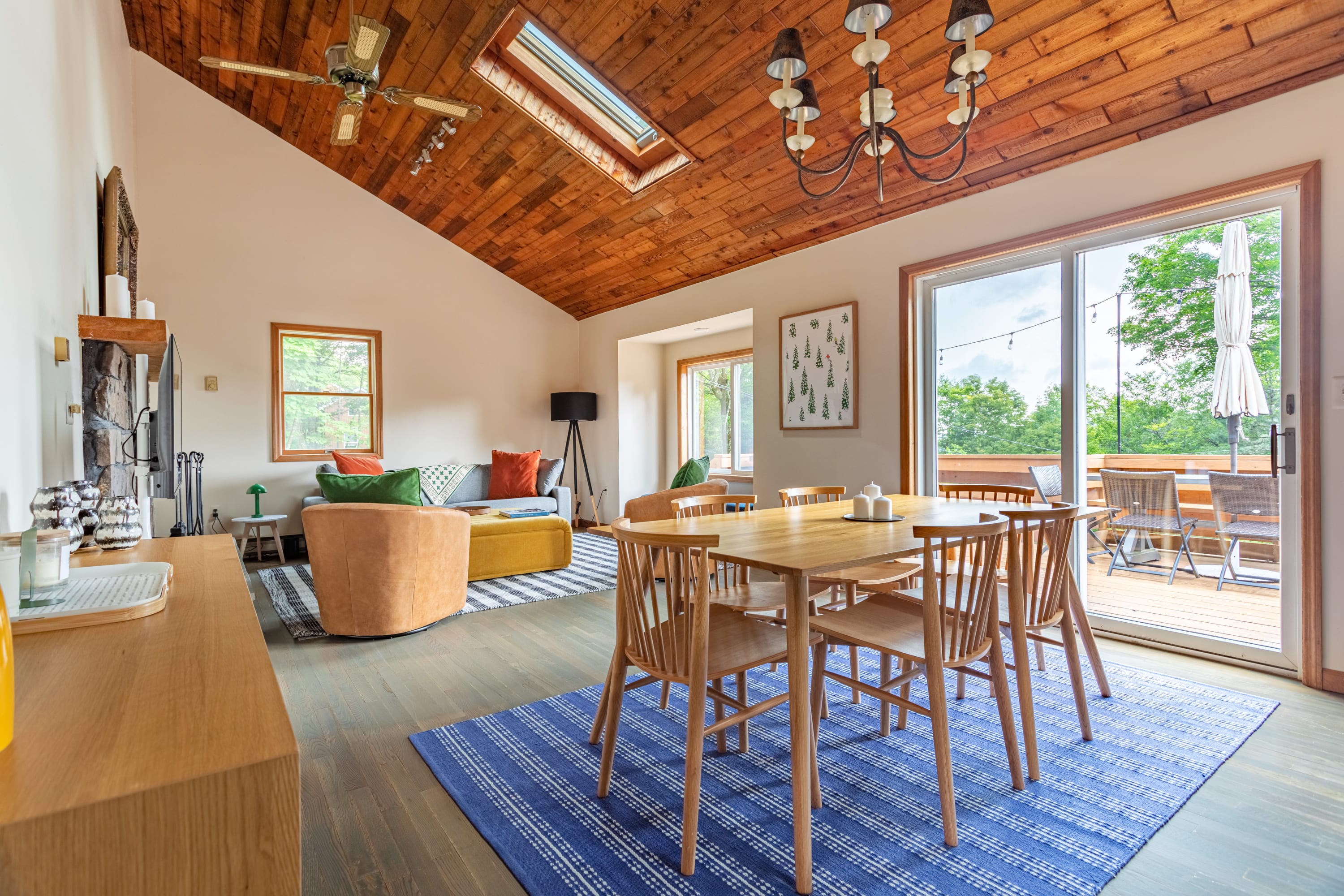 Open-concept living and dining area with a vaulted wooden plank ceiling and skylight. A wooden dining table with chairs sits on a blue rug, adjacent to a living space with a sofa and armchair, and a sliding glass door leads to a deck.