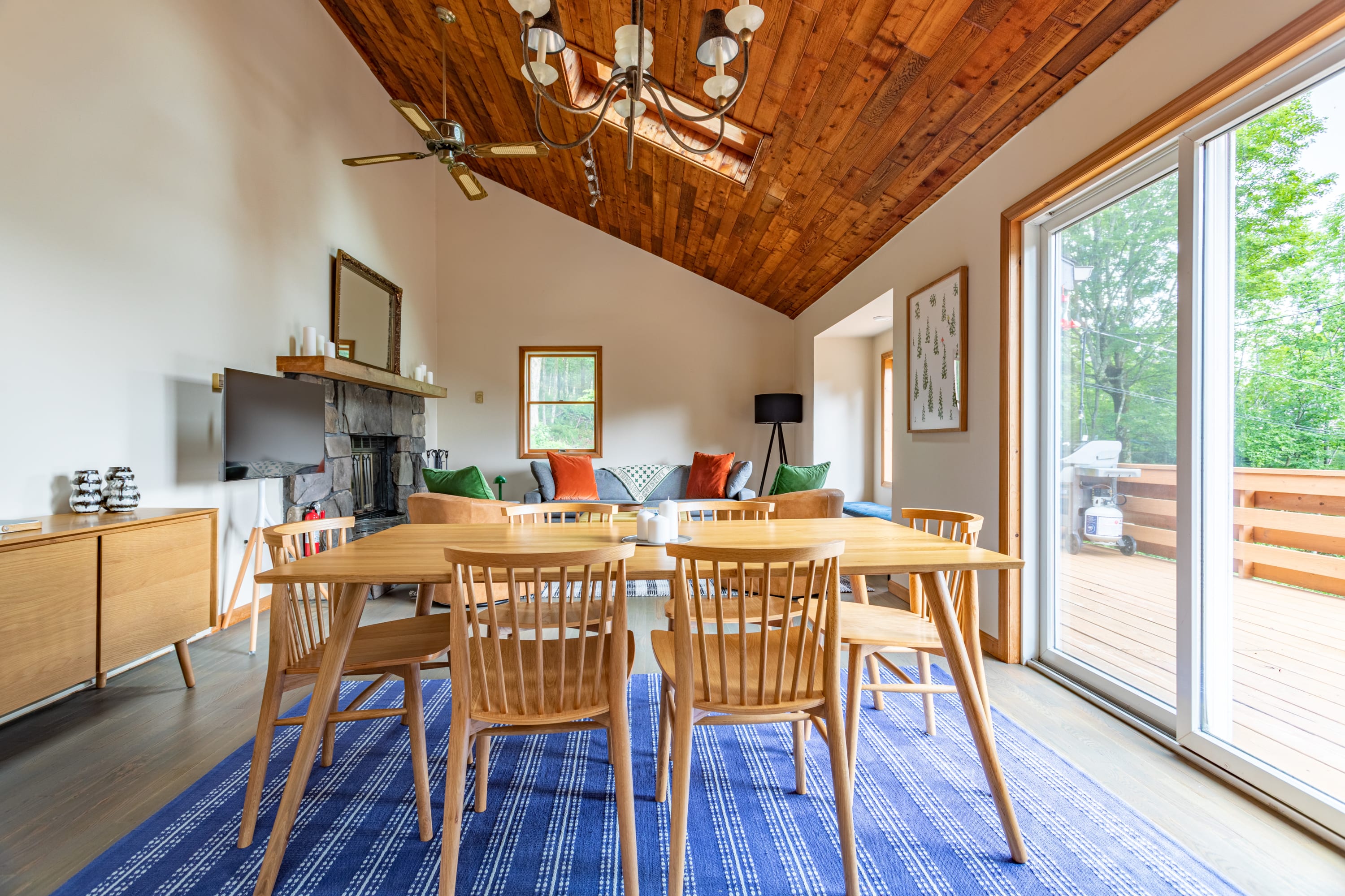 Open concept dining area featuring a light wood dining table with six matching chairs on a blue and white striped rug, under a vaulted wood-paneled ceiling with a chandelier and ceiling fan.