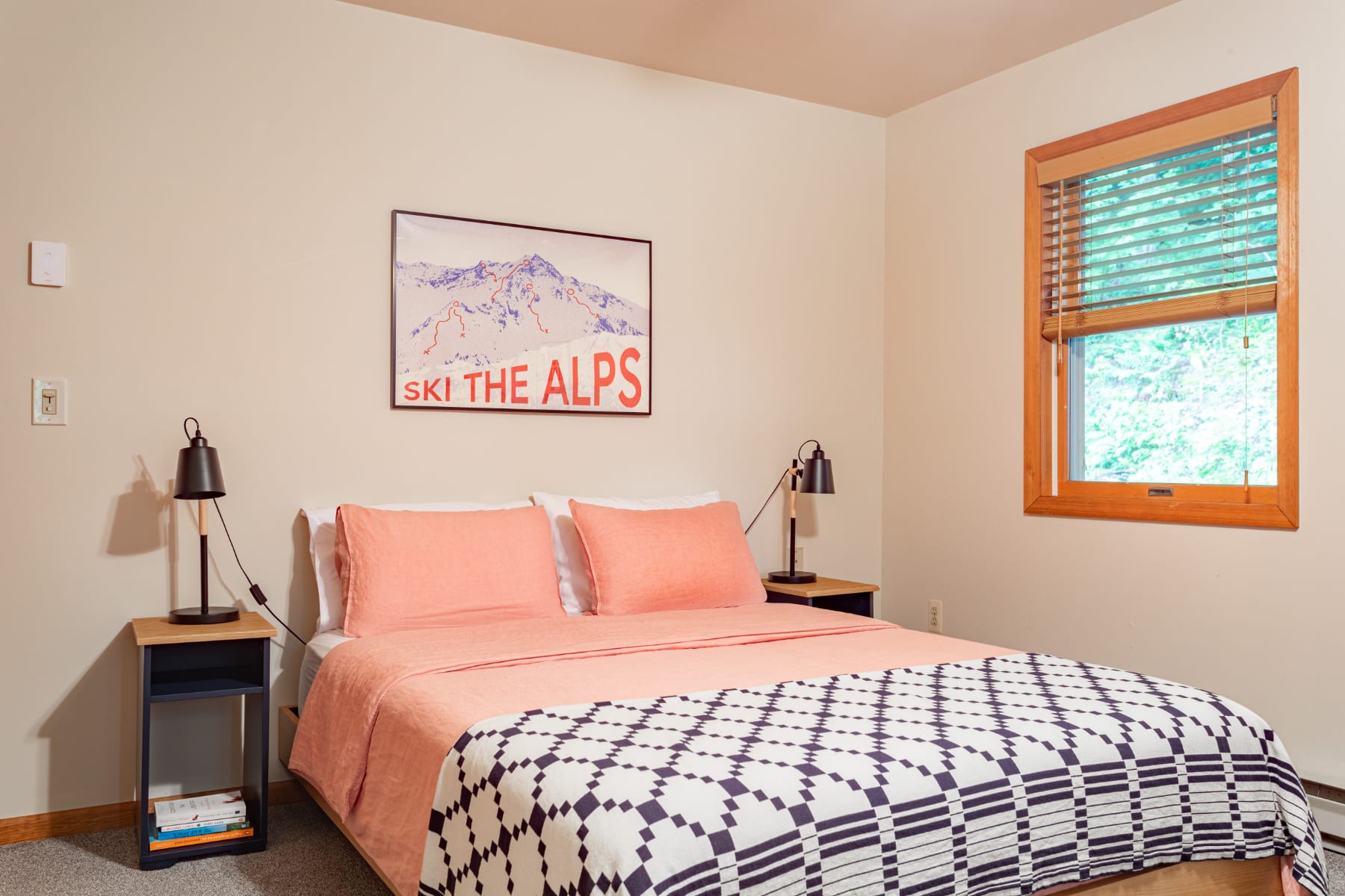 Cozy bedroom with a queen-sized bed featuring a patterned duvet and peach bedding, flanked by two black nightstands with lamps, and a "Ski The Alps" poster above the headboard.