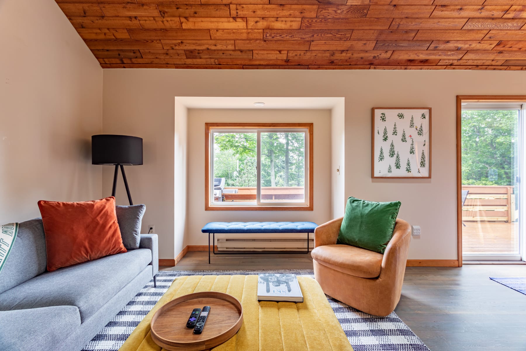 Bright living room with a gray sofa, yellow ottoman, and tan armchair on a striped rug, featuring a window nook with a blue bench and views of lush greenery outside.