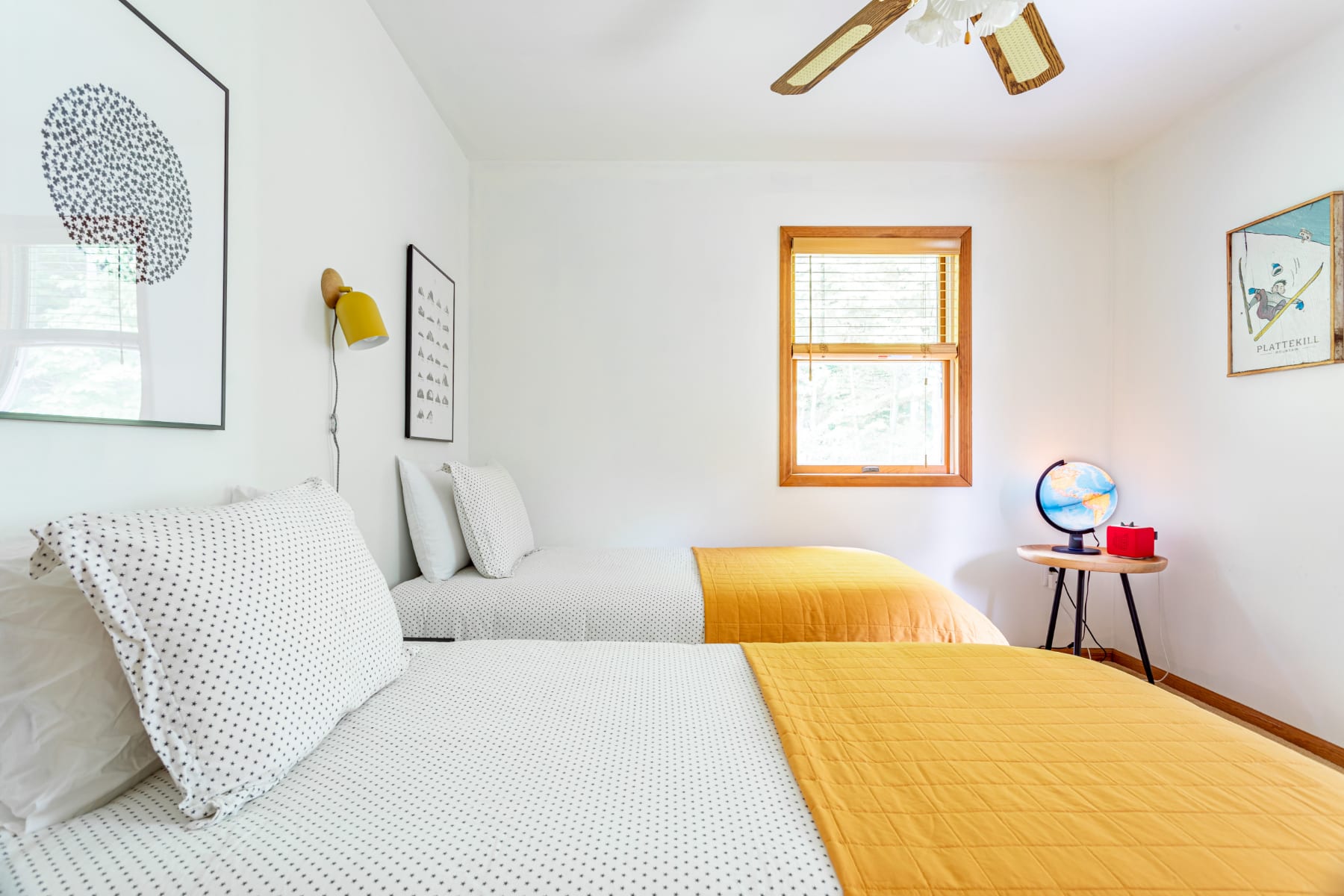 A bright bedroom with two twin beds, each covered with white polka-dot bedding and a yellow quilted throw. A small round side table with a globe sits between the beds, and a yellow wall sconce adds a pop of color.