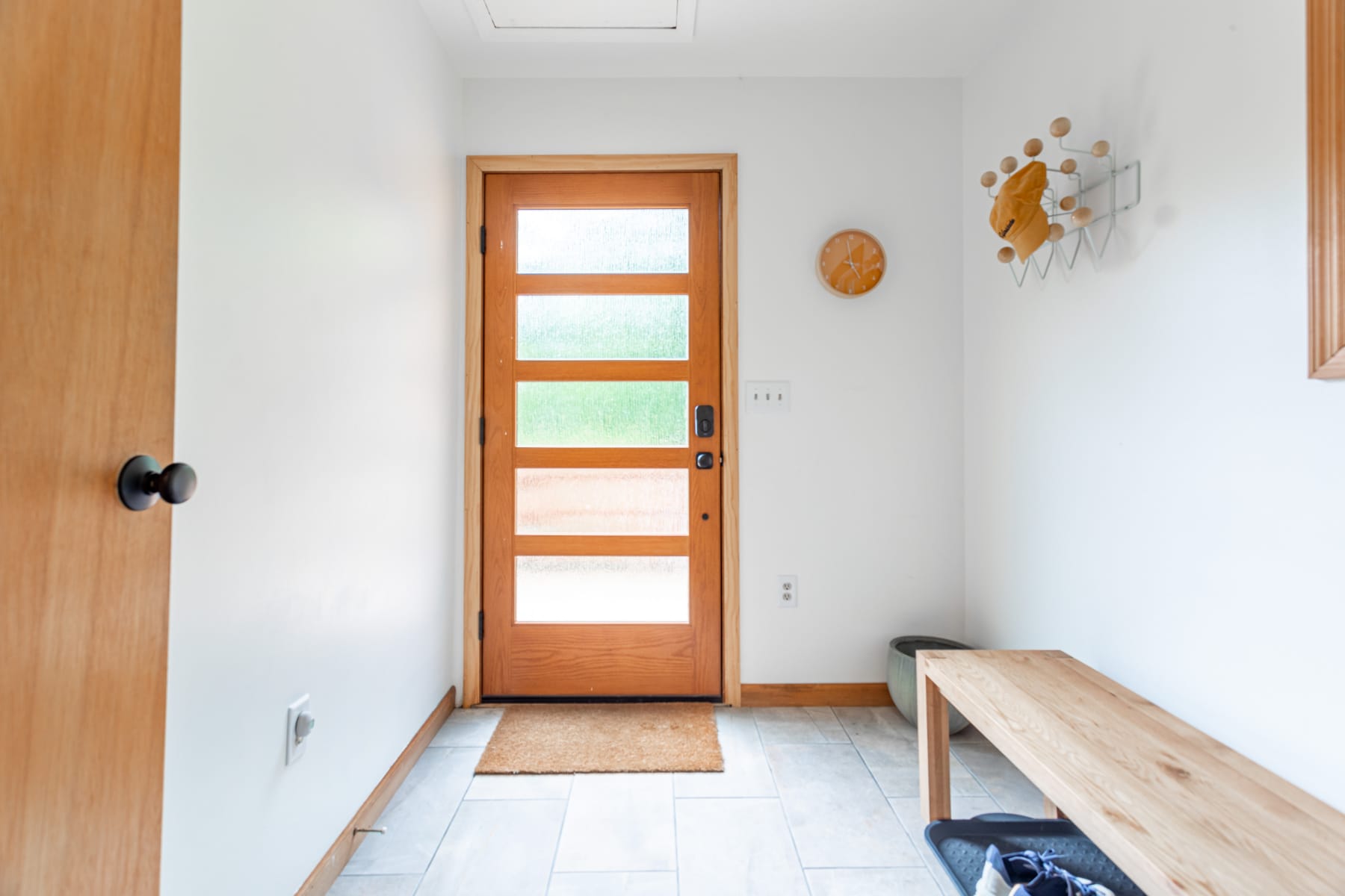Bright foyer with a light wood-framed glass panel door, white walls, light tile flooring, a wooden bench, and a modern coat rack with hats.