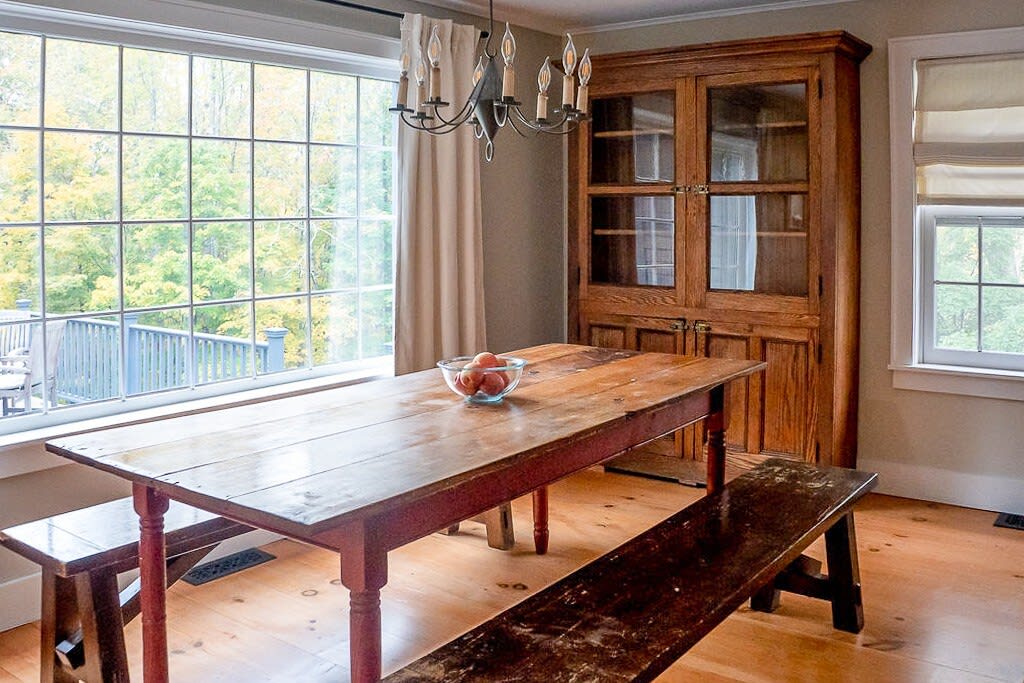 Rustic dining area with a long wooden farm table and benches, illuminated by a chandelier and natural light from a large window overlooking a deck and autumn trees. A tall wooden display cabinet is visible.