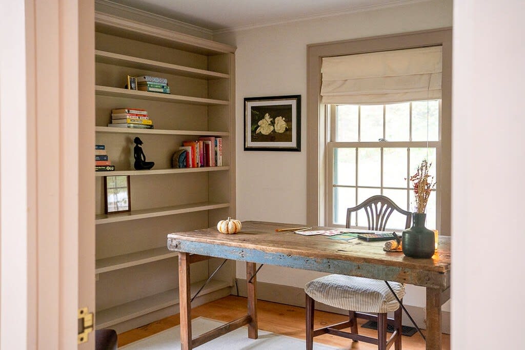 A cozy home office with a rustic wooden desk, a striped stool, and built-in bookshelves filled with books and decor. A window with a roman shade offers natural light, and a small pumpkin sits on the desk.