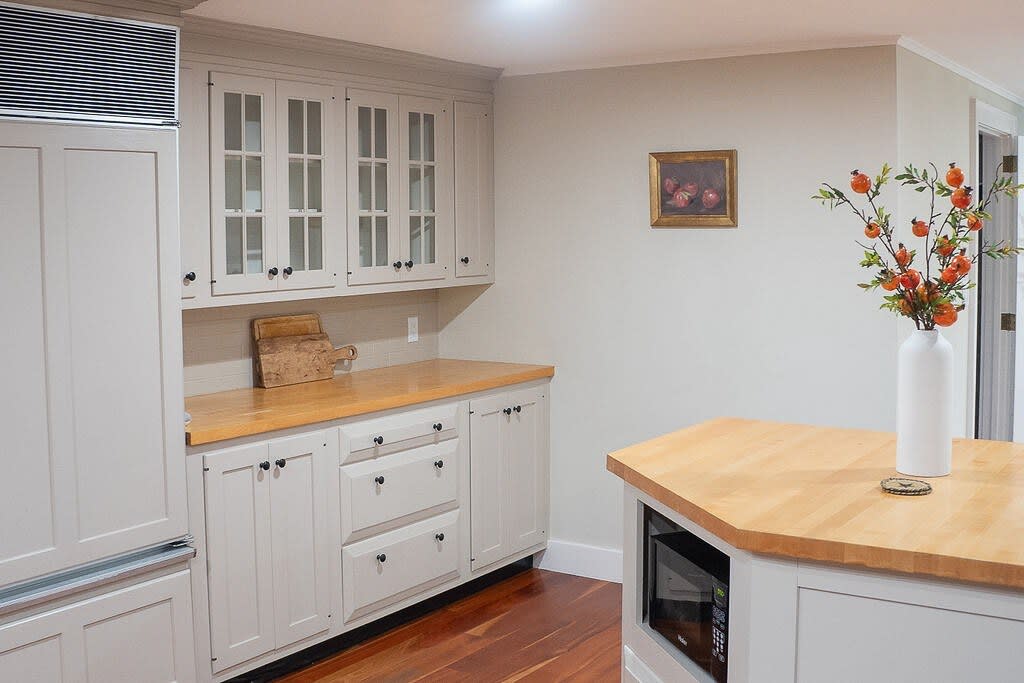 A bright kitchen featuring light gray shaker cabinets with black hardware, light wood countertops, and a built-in microwave in a kitchen island. A vase with decorative branches adds a touch of color.