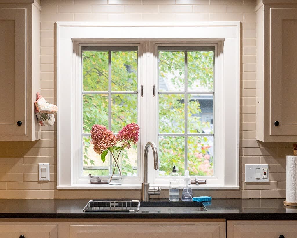 A bright kitchen sink area with a white framed window overlooking green trees, featuring a stainless steel faucet, black countertop, and light beige subway tile backsplash. A vase with pink flowers sits on the windowsill.