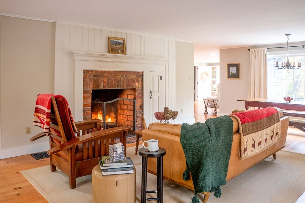 Cozy living room with light walls, wide-plank wood floors, and a large brick fireplace with a white mantel. A tan leather sofa and a wooden armchair with colorful throws create a warm seating area.