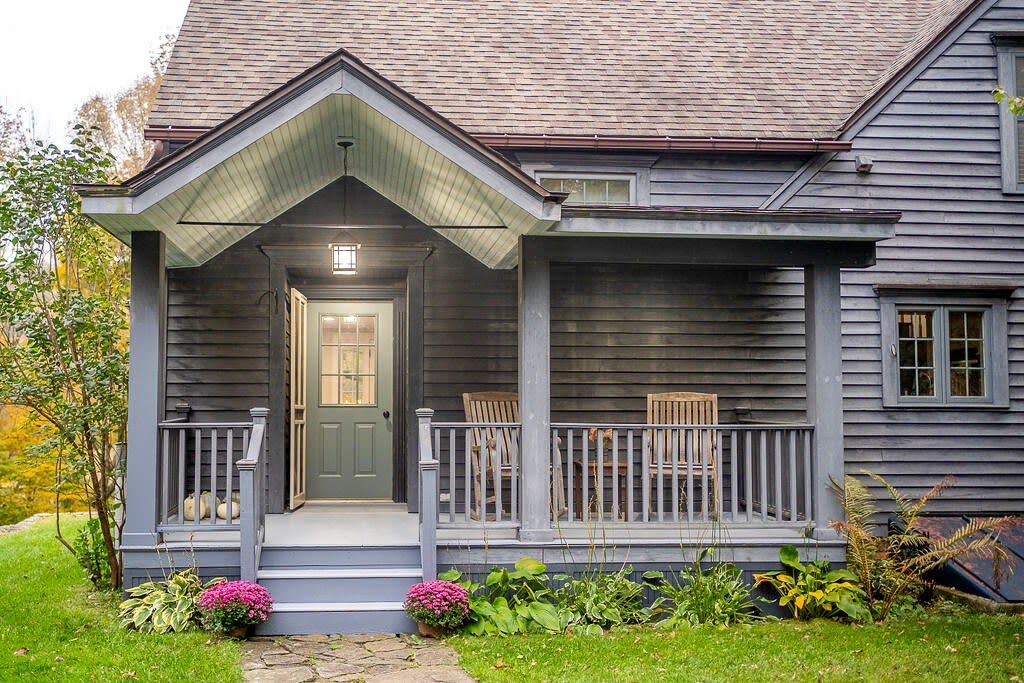 The inviting front porch of a dark grey cabin, featuring a light green front door and two wooden chairs on the porch. Lush green landscaping with flowering plants surrounds the entrance.