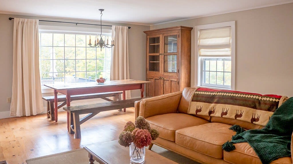 Open concept living and dining area featuring a tan leather sofa with a green throw, and a rustic wooden dining table with benches. A large window offers views of the outdoors, and a wooden display cabinet stands against the wall.