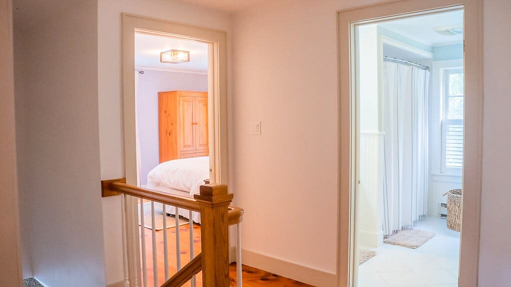 An upstairs hallway with light-colored walls and wood floors, showing doorways leading to a bedroom with a wooden wardrobe and a bathroom with a shower curtain.