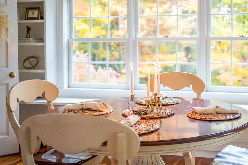 Close-up of a round wooden dining table set with placemats and candles, positioned in front of a large window offering a bright view of colorful autumn trees. Cream-colored dining chairs are partially visible.