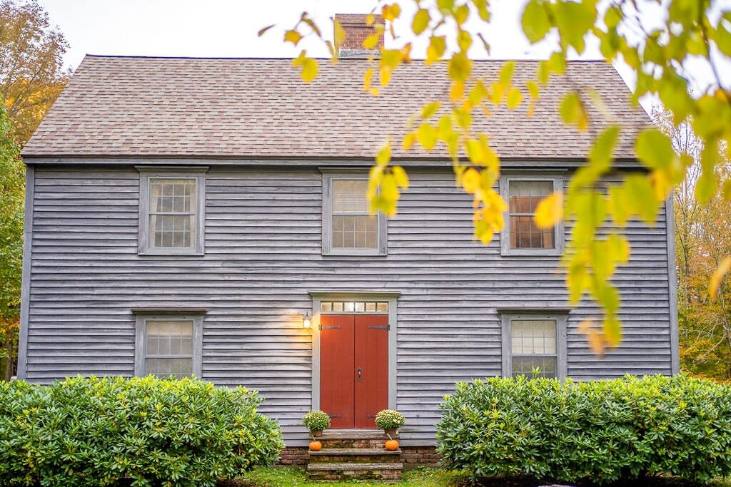 A classic two-story gray clapboard house with a brown shingle roof and a striking red double door at its center. Lush green bushes line the front, while vibrant autumn leaves frame the top of the scene.