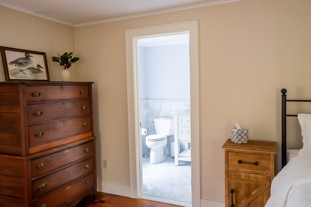 A close-up of a bedroom corner showing a tall wooden dresser with a framed picture and plant, and an open doorway leading into a light-tiled bathroom with a toilet and vanity visible.
