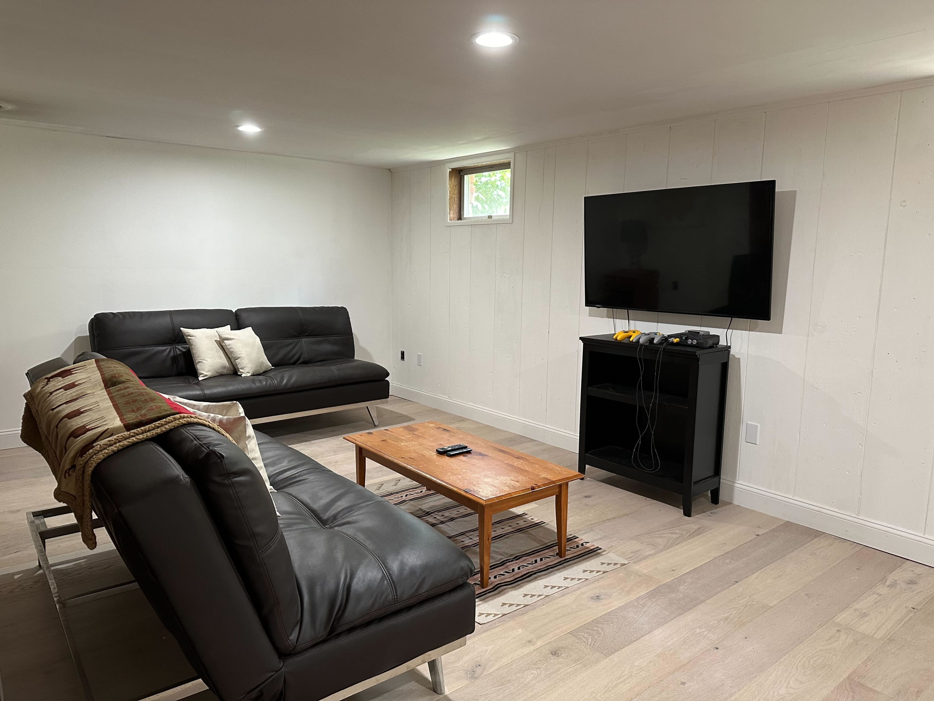 A modern living space with two black faux leather sofas facing a wall-mounted television above a black media console. A wooden coffee table sits on a patterned rug, and the walls are white shiplap.