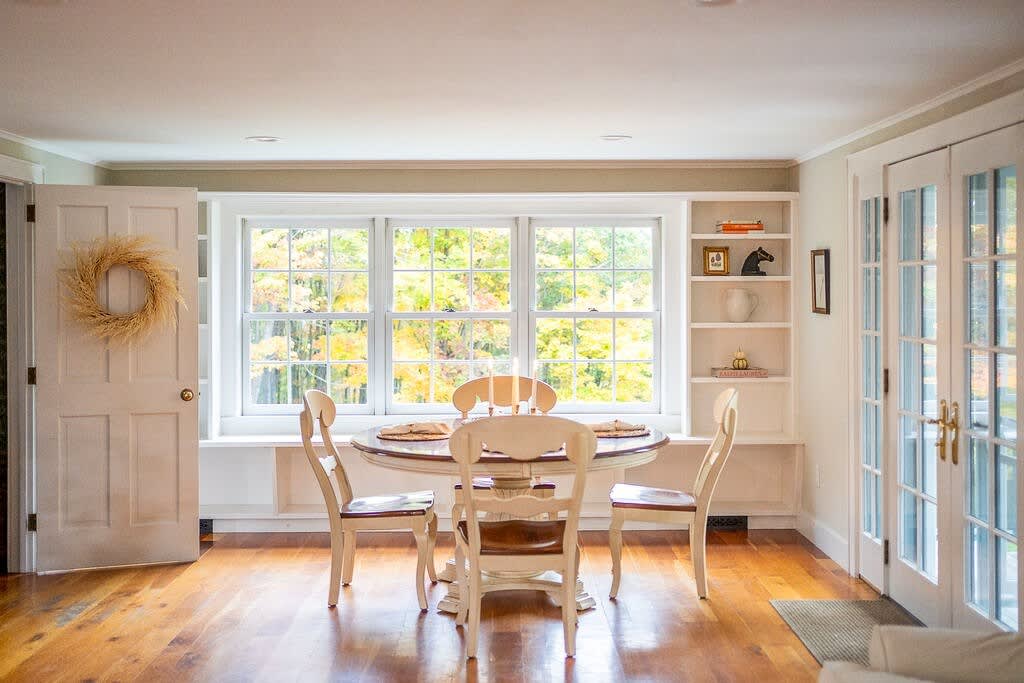 Bright dining area with a round wooden table and four cream-colored chairs, set against a large window overlooking vibrant autumn trees. Built-in shelves flank the window, and French doors lead to another room.