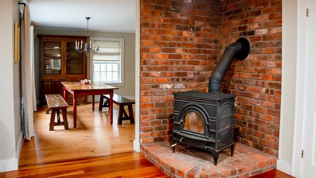 A rustic dining area featuring a black cast iron wood stove on a brick hearth, set against a red brick accent wall. Beyond, a wooden dining table with benches and a hutch are visible, all on warm-toned hardwood floors.