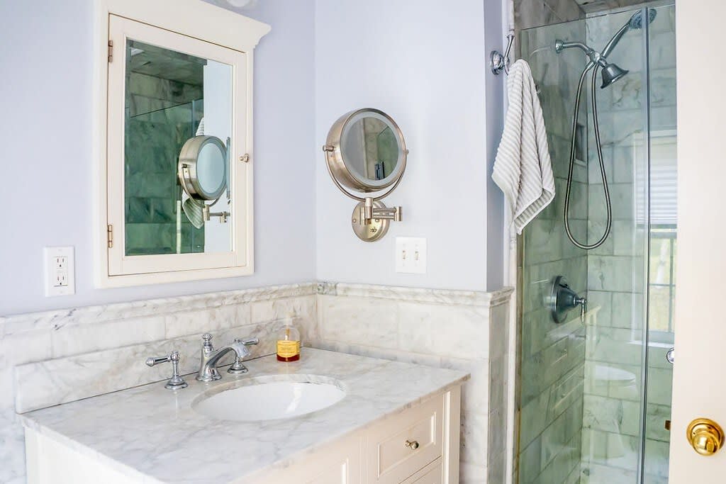 A modern bathroom with a white marble-topped vanity, an undermount sink, and chrome fixtures. A glass-enclosed shower with light green tiled walls is visible next to a wall-mounted magnifying mirror.