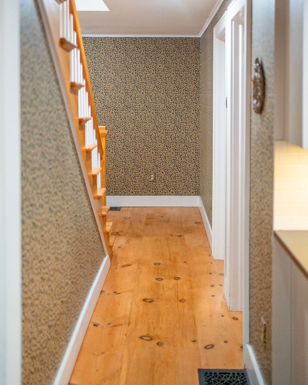 A narrow hallway or foyer with patterned wallpaper and light wood floors, leading towards a staircase with white balusters and a wooden handrail.