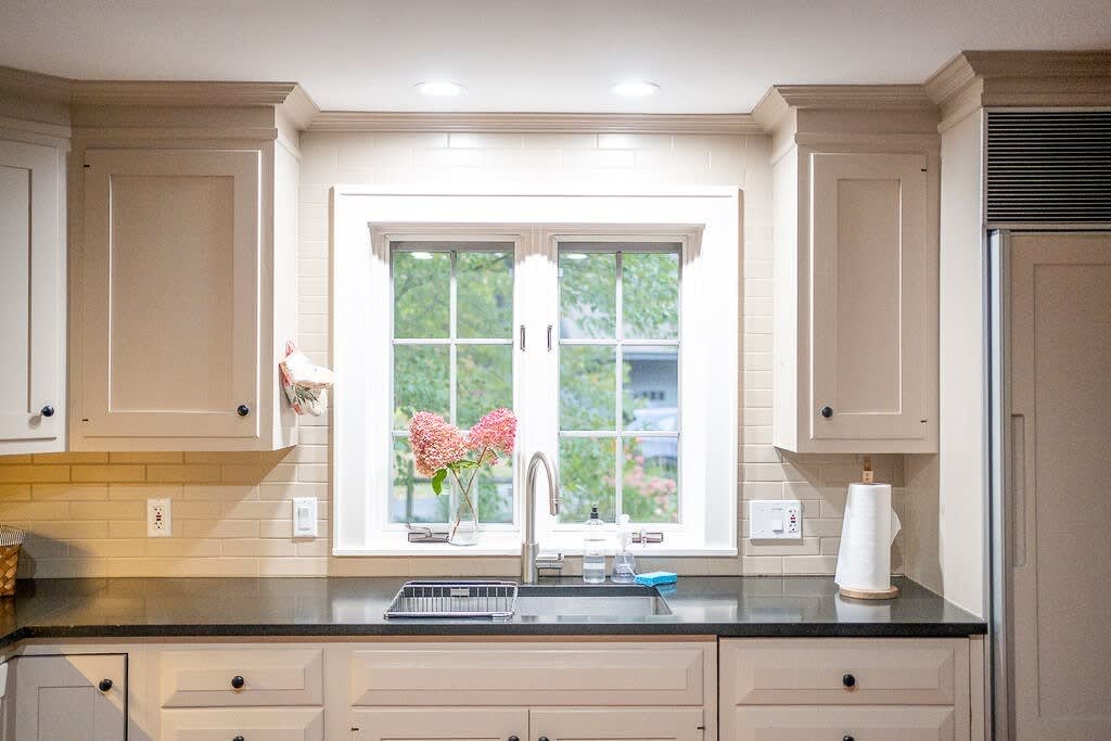 A well-lit kitchen with white shaker cabinets, a black countertop, and a window above the sink looking out to trees. The sink area includes a stainless steel faucet and a light beige subway tile backsplash.