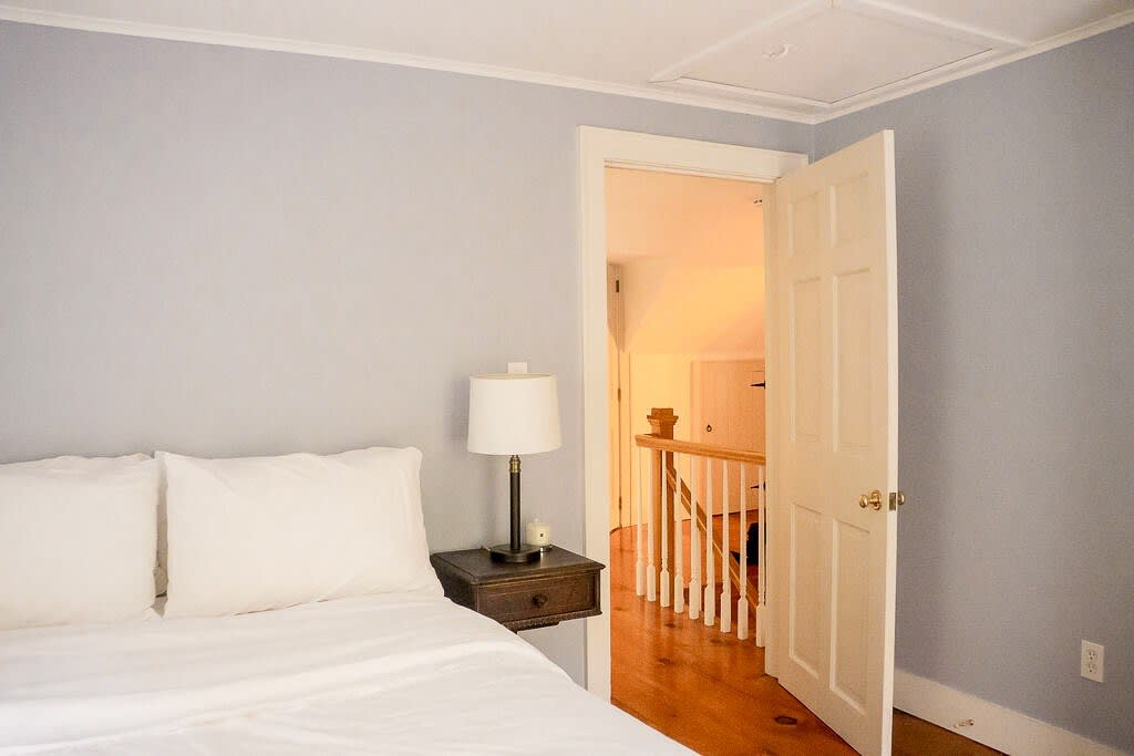 A view of a bedroom with light blue walls and a bed with white bedding, featuring a dark wooden floating bedside table with a lamp. An open door reveals a hallway with a wooden staircase and railing.