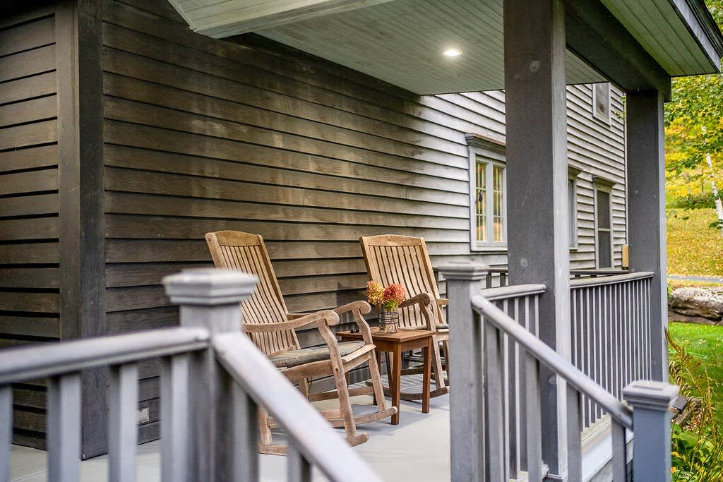 A cozy wooden porch with dark gray siding, featuring two rustic wooden rocking chairs and a small side table with a vase of flowers, overlooking a lush green and autumn-colored landscape.