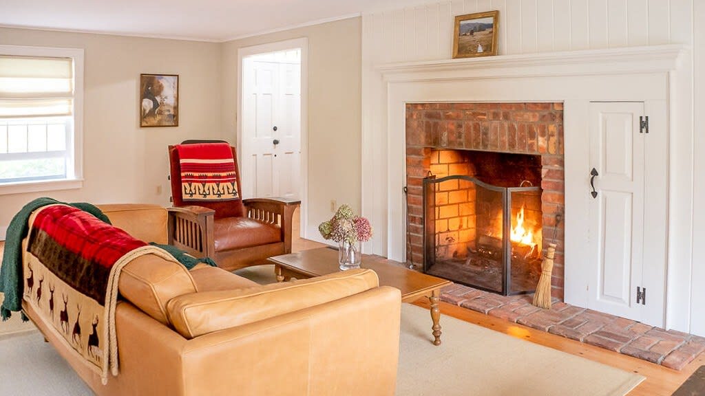 A warm living room with a brick fireplace, complete with a fire burning and a metal screen, flanked by a white built-in cabinet. A light brown leather sofa with a patterned throw blanket and a matching armchair are arranged around a wooden coffee table.