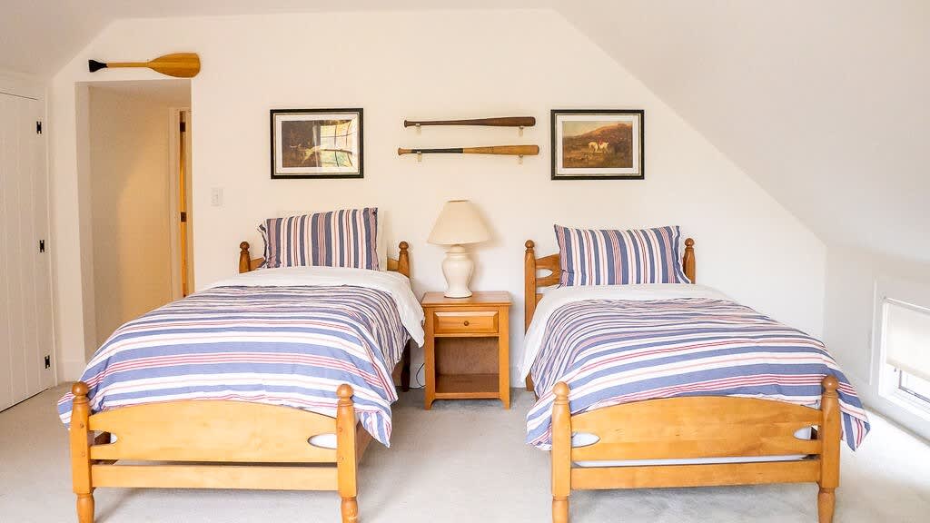 A wider view of the attic bedroom with two twin beds, showing a wooden nightstand between them with a lamp. The room has white walls, a sloped ceiling, and decorative elements like framed art and wooden oars.