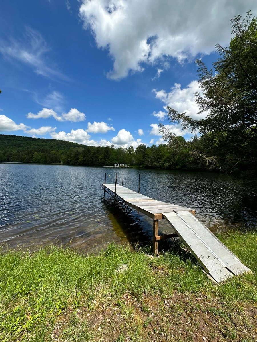 A wooden dock extends into a calm lake with clear, shallow water, surrounded by lush green grass and distant tree-covered hills under a bright blue sky with fluffy white clouds.