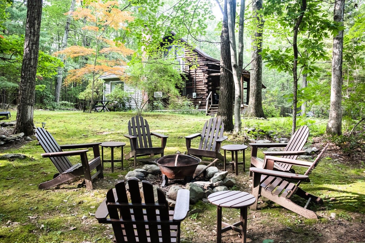 Outdoor fire pit area surrounded by six wooden Adirondack chairs and small side tables, set on a grassy lawn amidst a dense forest. The log cabin is visible in the background.