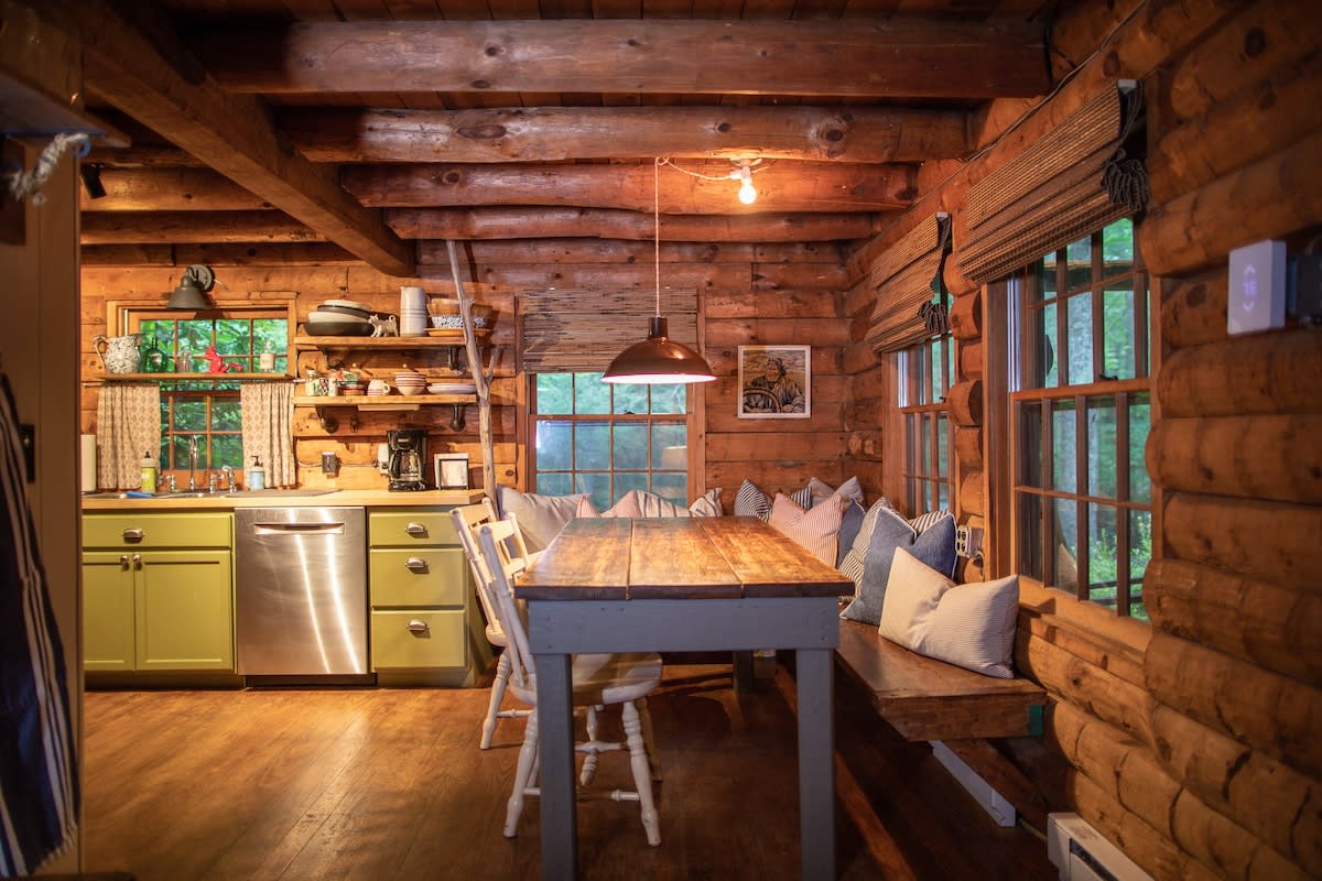 Rustic dining area with a long wooden table and bench seating, featuring log walls and wood-beamed ceilings. The adjacent kitchen has light green cabinets and stainless steel appliances.