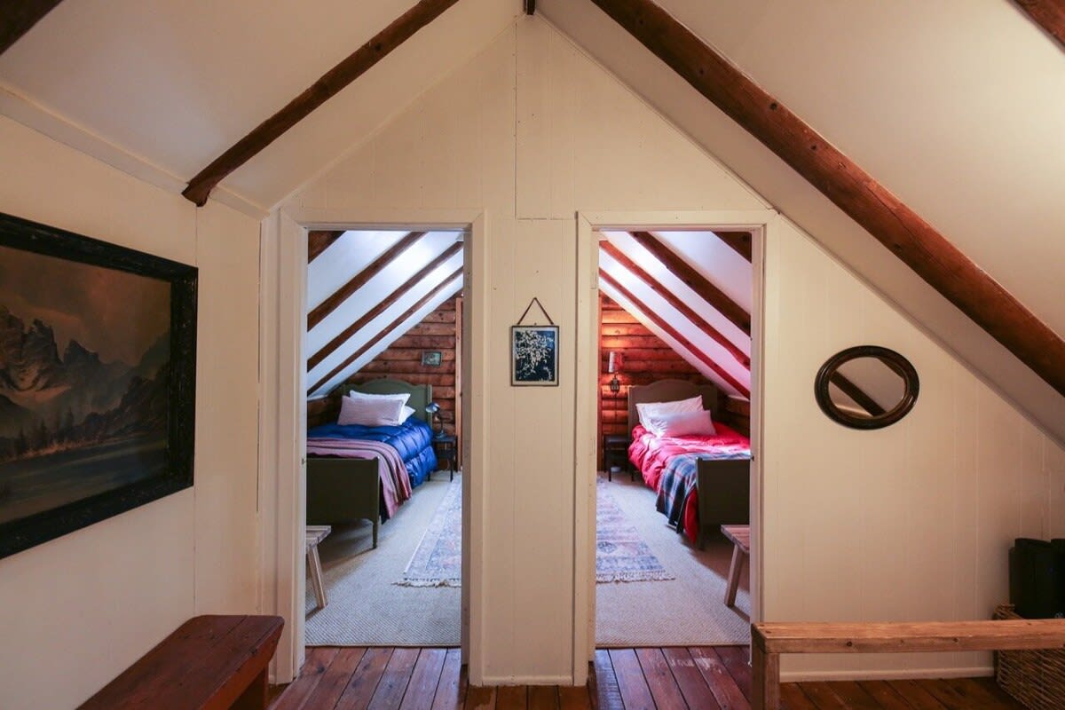 Attic landing area with white walls and exposed wood beams, featuring two doorways leading to separate bedrooms. The floor is light wood, and a large framed artwork hangs on the left wall.