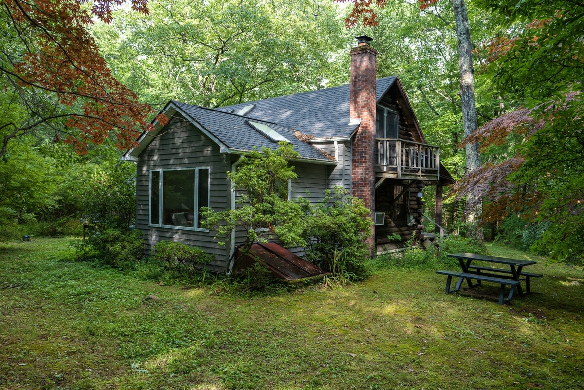 A charming cabin with a mix of log and gray siding, surrounded by a vibrant green lawn and mature trees. A brick chimney and a small balcony are visible on the log section, with a black picnic table in the foreground.