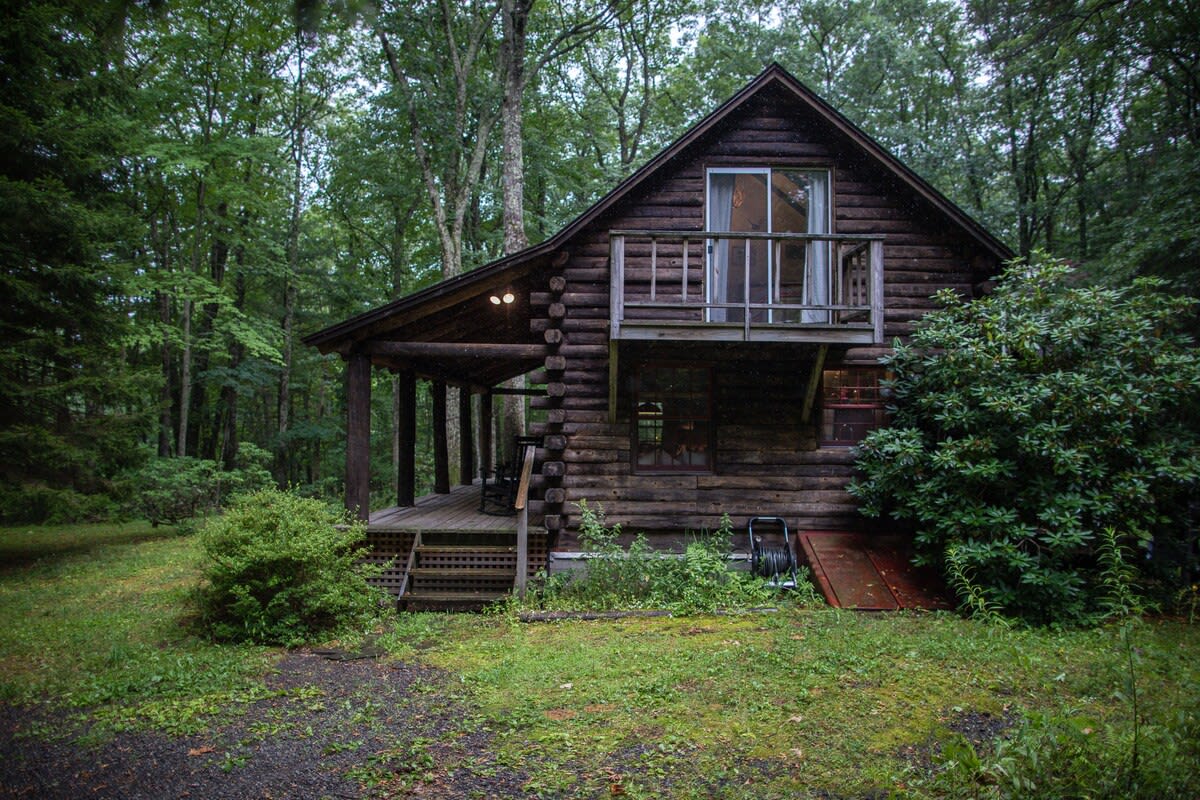 The front view of a rustic log cabin surrounded by dense green trees, featuring a small balcony on the second floor and a covered porch below. A wooden staircase leads up to the porch, and a red cellar door is visible on the right.