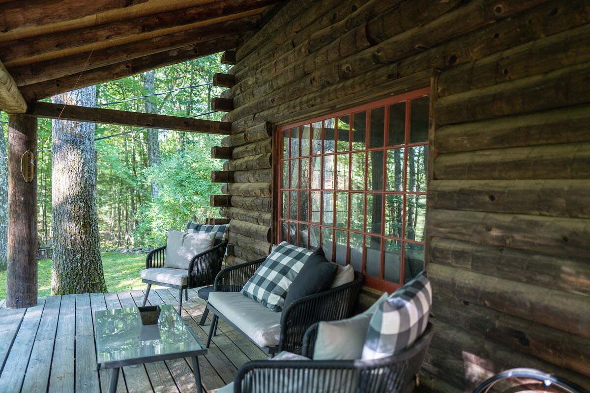 Covered wooden porch with comfortable outdoor lounge seating, including a sofa and armchairs with plaid cushions, set against rustic log walls. The area offers a serene view of the surrounding forest.